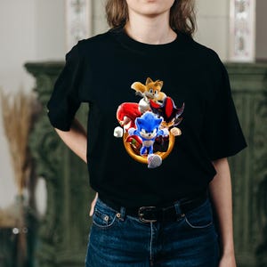 May include: Black t-shirt featuring a colorful graphic of Sonic the Hedgehog and other characters. The design includes Sonic, Tails, Knuckles, and Shadow, with a gold ring. The shirt is made of a soft material.