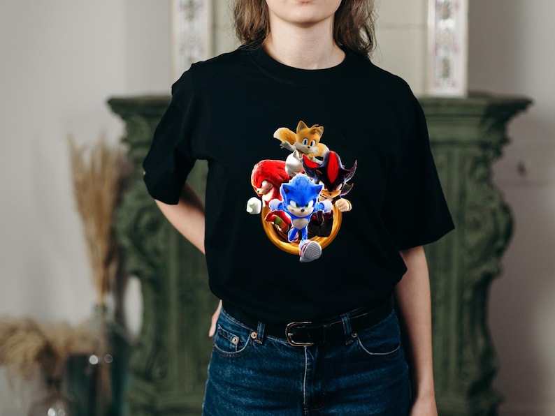 May include: Black t-shirt featuring a colorful graphic of Sonic the Hedgehog and other characters. The design includes Sonic, Tails, Knuckles, and Shadow, with a gold ring. The shirt is made of a soft material.