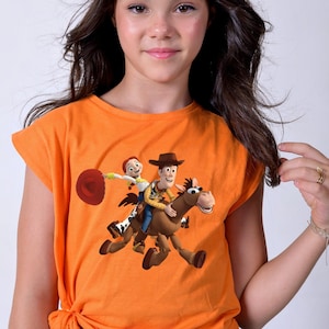 May include: Orange t-shirt featuring a graphic of Woody and Jessie from Toy Story riding a horse. The shirt has short sleeves and a knotted hem. The design is colorful and vibrant, perfect for fans of the animated movie.