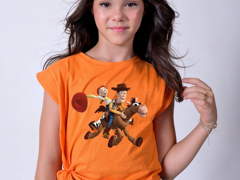 May include: Orange t-shirt featuring a graphic of Woody and Jessie from Toy Story riding a horse. The shirt has short sleeves and a knotted hem. The design is colorful and vibrant, perfect for fans of the animated movie.