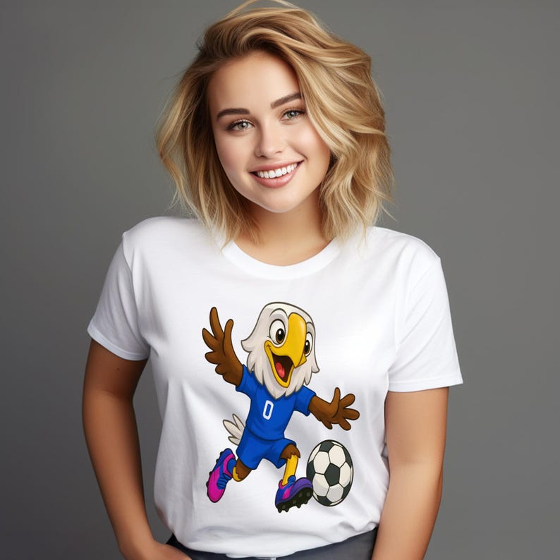 May include: A white short-sleeved t-shirt featuring a cartoon graphic of a bald eagle wearing a blue soccer jersey with the number zero, brown wings, and colorful cleats, shown mid-action while kicking a black and white soccer ball.
