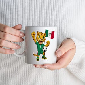 May include: A white ceramic mug featuring a cartoon jaguar wearing a green soccer jersey with the number 8, holding a soccer ball, and standing next to a waving Mexican flag. The design is centered on the mug, showcasing vibrant colors.