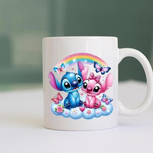 May include: White ceramic mug with a colourful illustration of two cartoon characters, one blue and one pink, on a cloud under a rainbow. Butterflies and flowers decorate the design.
