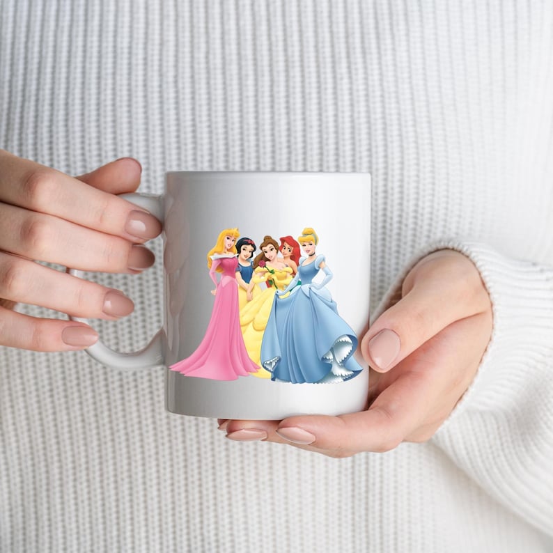 May include: White ceramic mug with a design of animated princesses in various gowns. The princesses are depicted in pink, yellow, and blue dresses. The mug has a white handle and is held by a person wearing a white sweater.