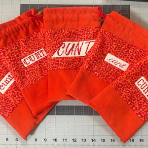 May include: A collection of orange drawstring bags. Each bag features a leopard print panel with the word "cunt" embroidered on it. The bags are arranged on a gray cutting mat.