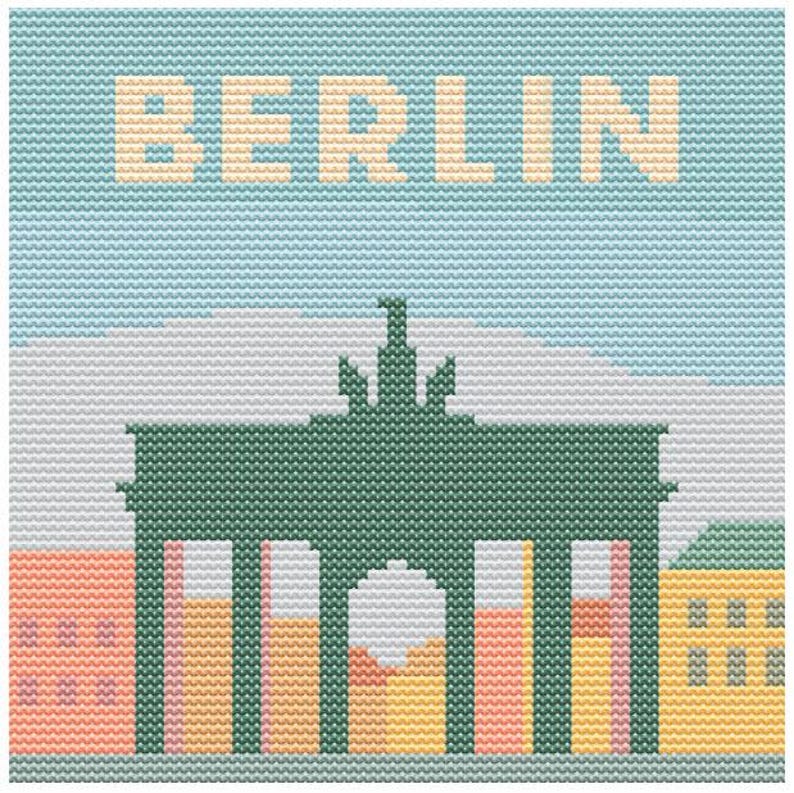 May include: Cross-stitch design featuring the word "BERLIN" above a green Brandenburg Gate illustration. The sky is light blue, and the buildings below are in shades of orange, yellow, and pink.