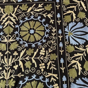 May include: Black textile with intricate embroidery featuring floral and leaf patterns in shades of blue, green, and cream. The design includes stylized flowers and scrolling vines, creating a detailed and ornate look. The overall aesthetic is reminiscent of traditional textile art.