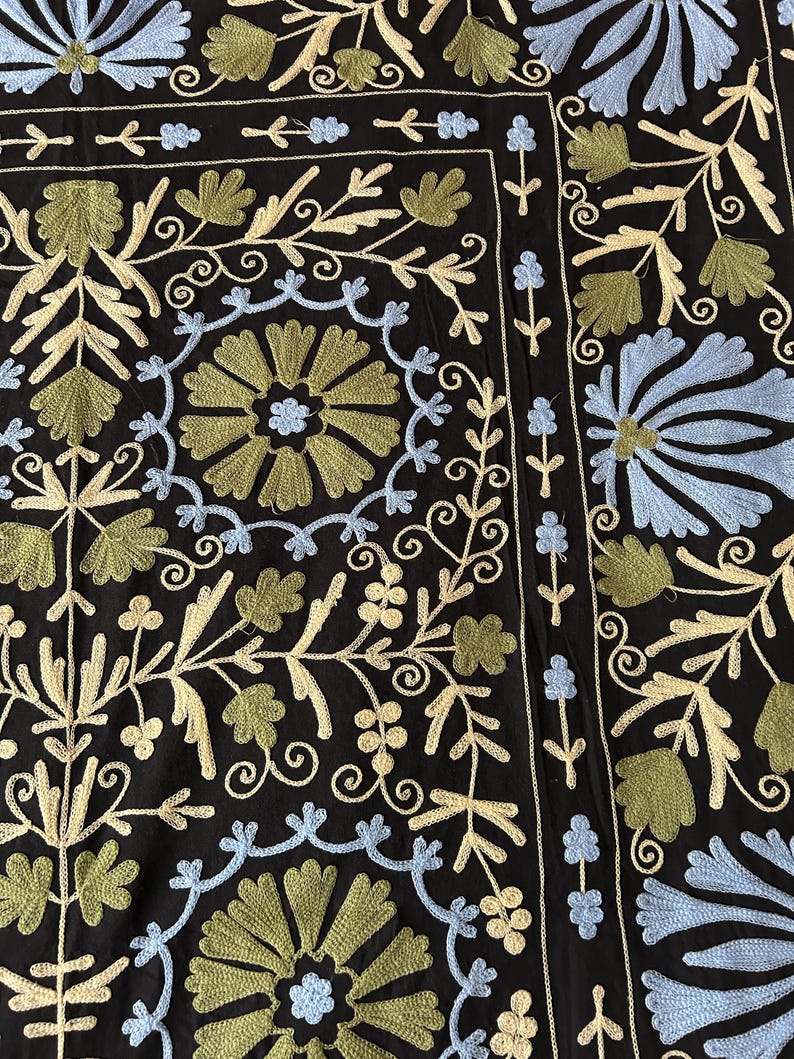 May include: Black textile with intricate embroidery featuring floral and leaf patterns in shades of blue, green, and cream. The design includes stylized flowers and scrolling vines, creating a detailed and ornate look. The overall aesthetic is reminiscent of traditional textile art.
