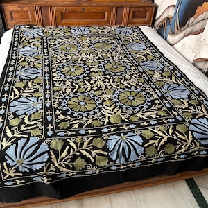 May include: Black embroidered bedspread with a floral design in shades of blue, green, and cream. The intricate pattern covers the entire surface, creating a rich and detailed textile. The bedspread is displayed on a wooden bed frame.