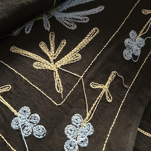 May include: Close-up of black fabric with detailed embroidery. The design features light blue and beige floral and leaf patterns, along with delicate white stitching. The embroidery creates a textured, artistic effect.