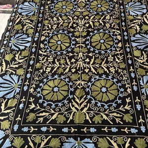 May include: Black textile with intricate floral embroidery in shades of blue, green, and cream. The design features stylized flowers and foliage, creating a symmetrical pattern. The edges are bordered with a similar design.