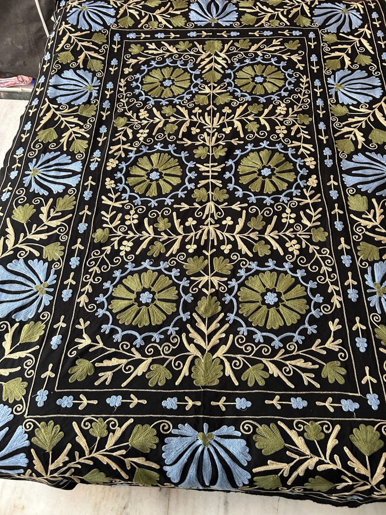 May include: Black textile with intricate floral embroidery in shades of blue, green, and cream. The design features stylized flowers and foliage, creating a symmetrical pattern. The edges are bordered with a similar design.