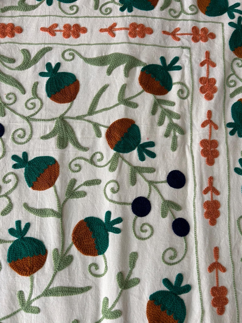 May include: Close-up of a white textile with embroidered floral patterns. The design features green leaves and stems, with brown and green fruit-like shapes. Orange floral accents and dark blue circular details complete the design.