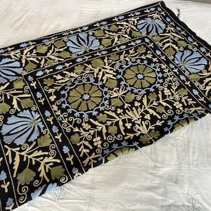 May include: Black textile with intricate floral embroidery in shades of blue, green, and cream. The design features stylized flowers and foliage, creating a decorative pattern. The textile is likely a decorative piece, such as a throw or wall hanging.