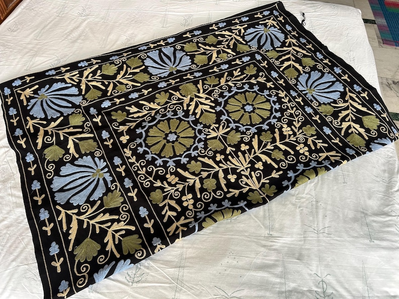May include: Black textile with intricate floral embroidery in shades of blue, green, and cream. The design features stylized flowers and foliage, creating a decorative pattern. The textile is likely a decorative piece, such as a throw or wall hanging.