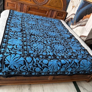 May include: A black blanket with a detailed blue floral pattern. The blanket is spread across a wooden bed frame. The floral design is intricate and covers the entire surface of the blanket, creating a visually rich textile.