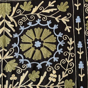 May include: Black textile with intricate embroidery. The design features floral and leaf patterns in shades of green, light blue, and cream. The embroidery is detailed and covers the surface, creating a textured appearance.