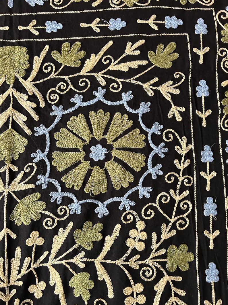 May include: Black textile with intricate embroidery. The design features floral and leaf patterns in shades of green, light blue, and cream. The embroidery is detailed and covers the surface, creating a textured appearance.
