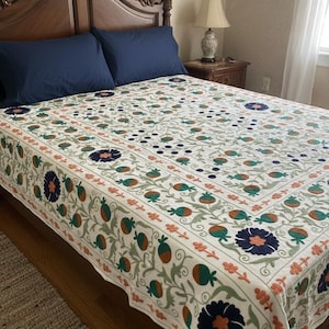 May include: Cream-coloured bedspread with an embroidered floral pattern in shades of green, orange, and navy blue. The bedspread is on a wooden bed frame with two navy blue pillows. The room has a neutral colour scheme.
