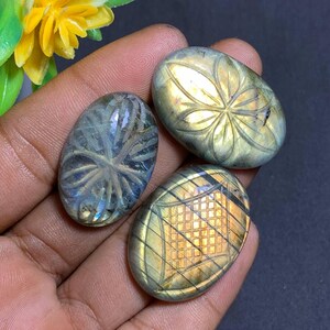 May include: Three oval-shaped labradorite stones with carved designs. One stone features a floral pattern, another a geometric grid, and the third a stylized sunburst. The stones have iridescent blue, green, and gold hues.