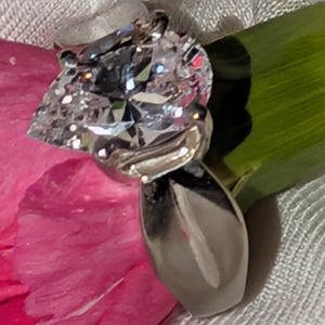 May include: A silver-toned ring featuring a large, pear-shaped clear faceted stone set in a polished metal band. The ring is positioned against a vibrant pink petal and a bright green stem, highlighting the reflective surfaces of the stone.
