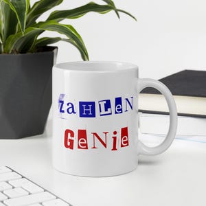 Puede incluir: Taza de cerámica blanca con las palabras "Zahlen Genie" impresas en letras de bloque azules y rojas. La taza está sobre una superficie blanca con una planta y un teclado en el fondo.