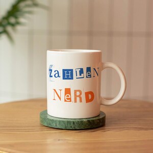 May include: White ceramic mug with the word "Zahlen Nerd" printed in a cut-out style with blue and orange letters. The mug sits on a green coaster on a wooden table.