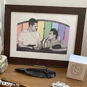 Puede incluir: Un dibujo en blanco y negro enmarcado de dos hombres en una cama con una cabecera arcoíris. El dibujo está sobre una mesa de madera con una escultura de búho blanca, una pequeña pala negra, un objeto decorativo dorado y blanco, una escultura de árbol blanca y una caja blanca con el texto "BAIES" en ella.
