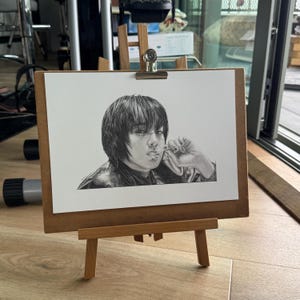 May include: A pencil portrait of a person with dark hair and a leather jacket, holding a lollipop. The artwork is displayed on a small wooden easel with a clip. The drawing is in black and white.