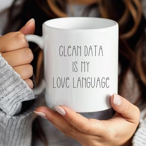 May include: White ceramic mug with the text "CLEAN DATA IS MY LOVE LANGUAGE" in black lettering. The mug has a standard handle and a cylindrical shape. The mug is held by a person wearing a gray sweater.