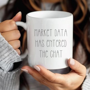 May include: White ceramic mug with the text "MARKET DATA HAS ENTERED THE CHAT" in black, held by a person. The mug has a curved handle and a cylindrical shape. The background is blurred.
