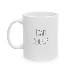 May include: White ceramic mug with the words "TEAM VLOOKUP" printed in black. The mug has a curved handle and a cylindrical shape. The text is in a simple, sans-serif font.