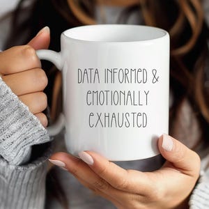 May include: White ceramic mug with the text "DATA INFORMED & EMOTIONALLY EXHAUSTED" in black lettering. The mug has a curved handle and is held by a person wearing a gray sweater. The mug is a standard size for coffee or tea.