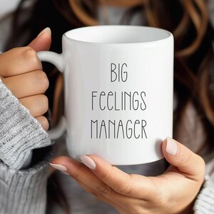May include: White ceramic mug with the text "BIG FEELINGS MANAGER" in black, sans-serif font. The mug has a handle and is held by a person wearing a gray sweater. The mug is cylindrical and appears to be empty.