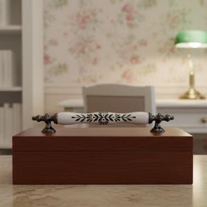 May include: A rectangular, dark brown wooden box with a decorative handle. The handle is white with black floral designs and dark metal ends. The box is on a light-colored surface.