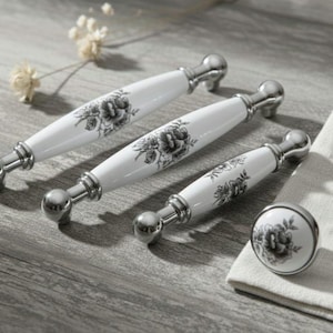 Puede incluir: Un juego de tiradores de armario y un pomo de cerámica blanca con un diseño floral en negro y gris. Los tiradores tienen una base de metal plateado. Los artículos están dispuestos sobre una superficie de madera con un paño blanco.