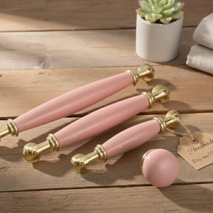 Op de afbeelding: Drie roze kastgrepen en een bijpassende knop met goudkleurige hardware. De grepen zijn in verschillende maten verkrijgbaar. Een klein label met het woord "Handmade" is aan de grepen bevestigd. Een kleine vetplant in een witte pot staat op de achtergrond.