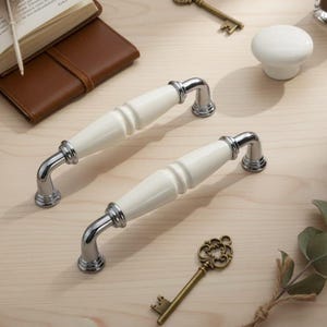 以下が含まれることがあります： クロム仕上げの金属アクセントが付いた、2つの白いセラミックキャビネットプル。プルは、湾曲したハンドルと装飾的なディテールを備えたクラシックなデザインです。白いセラミックノブ、ヴィンテージキー、革張りの本も表示されています。