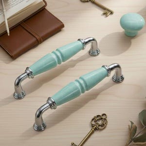 Può includere: Due maniglie per armadietti color acquamarina con dettagli argentati e un pomello coordinato sono esposte su una superficie in legno chiaro. Sono visibili anche una chiave vintage e un libro con copertina in pelle, che suggeriscono un tema vintage o antico.