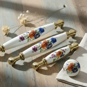 May include: A set of white ceramic cabinet pulls and a knob with a floral design in orange, purple, and blue. The hardware has gold-colored metal accents. The items are arranged on a light-colored wooden surface.