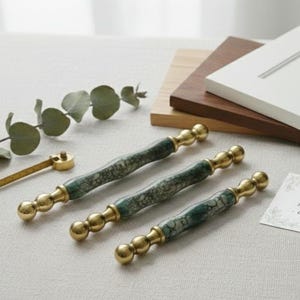 Peut inclure: Trois poignées de meubles avec un centre marbré vert et blanc et des extrémités dorées. Les poignées sont disposées sur une surface de couleur claire, avec une branche de verdure et un petit outil de mesure en arrière-plan.