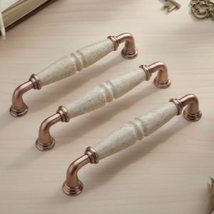May include: Three cabinet pulls with speckled cream-coloured centres and copper-coloured curved handles. The hardware is arranged on a light wood surface, suggesting a vintage or antique aesthetic.