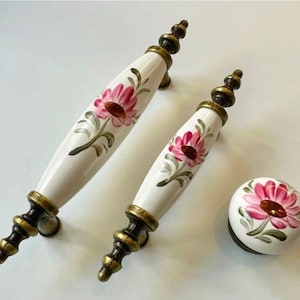 May include: A set of three decorative cabinet hardware pieces. Two white ceramic handles and a knob feature a pink floral design with green leaves. The hardware has bronze-colored metal accents.