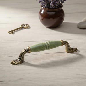 Peut inclure: Une poignée de meuble avec un centre en céramique craquelée verte et des extrémités en laiton antique. La poignée a un design incurvé et est posée sur une surface en bois clair. Une clé décorative et un petit vase sont en arrière-plan.