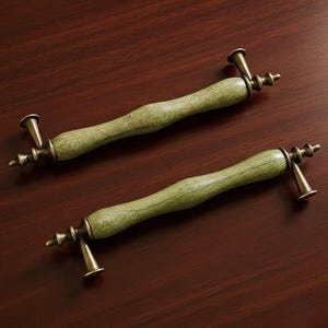 May include: Two decorative cabinet pulls with a light green, crackled finish and antique brass hardware. The pulls have a curved, ergonomic design and are set against a dark wood surface.