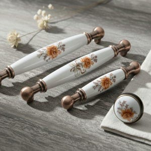 Puede incluir: Un juego de tiradores y un pomo de cerámica blanca con herrajes de color bronce. Cada pieza presenta un diseño floral en tonos naranja y marrón. El conjunto está dispuesto sobre una superficie de madera.