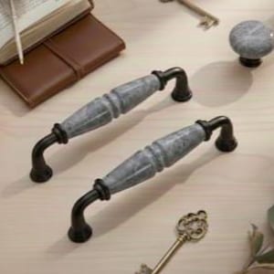 May include: Two gray marble-look cabinet handles with black curved supports are displayed on a light wood surface. A matching knob is visible, along with a leather wallet, open book, and decorative keys.