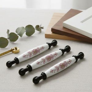 Op de afbeelding: Drie witte kastgrepen met bloemenmotief en zwarte uiteinden, samen met een gouden maatlepel, een takje eucalyptus en voorbeelden van kastdeuren. De grepen zijn ongeveer 15 cm lang.