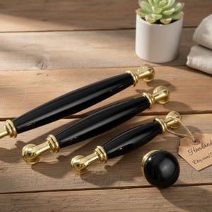 Pode incluir: Um conjunto de ferragens para armários pretas e douradas, incluindo três puxadores e um puxador redondo. Os puxadores têm um centro preto brilhante e detalhes dourados. As ferragens são exibidas em uma superfície de madeira.