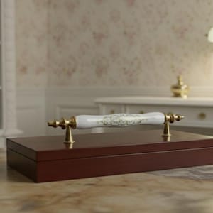 May include: A rectangular, dark brown wooden box with a decorative handle. The handle is white with a floral design and gold-colored metal accents. The box is on a light-colored surface.